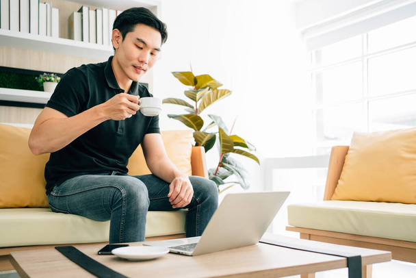 Homem asiático sentado e trabalhando no sofá enquanto bebe café. Empresário casual trabalhando no laptop em casa. Homem trabalhando em uma área de relaxamento no escritório. Período de quarentena comercial. - Foto, Imagem