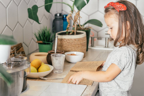 Chica morena de 3 años en diadema hornea tarta de manzana, se para cerca de la mesa con ingredientes, utensilios y libro de recetas. - Foto, imagen