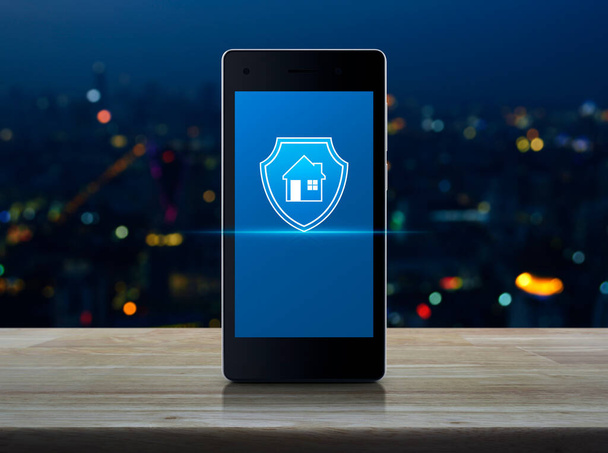 House with shield flat icon on modern smart mobile phone screen on wooden table over blur colorful night light city tower and skyscraper, Business home insurance and security online concept - Fotografie, Obrázek