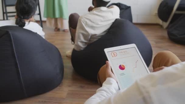 Tilting up of cropped man using tablet computer with business graphs on screen, sitting on black bean bag chair in coworking at daytime, Muslim Caucasian woman standing by chalkboard with diagram - Кадры, видео