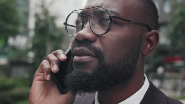 Ekstremalne zbliżenie ze śledzeniem afro-amerykańskiego biznesmena w okularach stojącego na zewnątrz i rozmawiającego przez telefon komórkowy - Materiał filmowy, wideo
