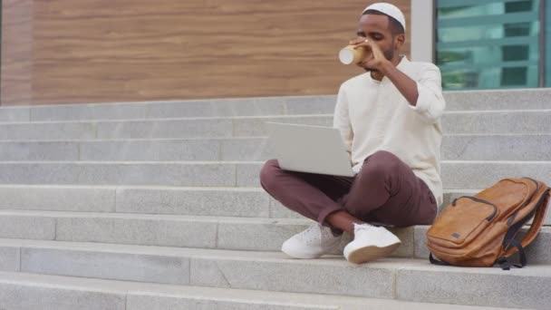 Πλήρης εικόνα του νεαρού μαύρου άνδρα που φοράει kufi, χρησιμοποιεί φορητό υπολογιστή, πίνει καφέ από χάρτινο κύπελλο, κάθεται σε σκαλοπάτια έξω από το κτίριο της πόλης την ημέρα - Πλάνα, βίντεο
