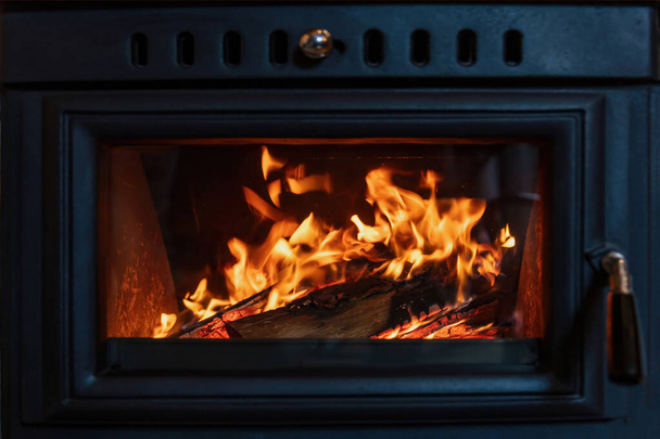 Дерев'яний пічний камін і палаючі дрова за скляними дверима, чорний метал, полум'я вогню випромінює тепло, теплий інтер'єр будинку
, - Фото, зображення