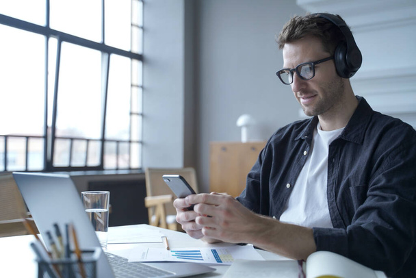 Német szabadúszó fickó fülhallgatóban ül a modern otthoni irodában laptopon, miközben okostelefont használ - Fotó, kép