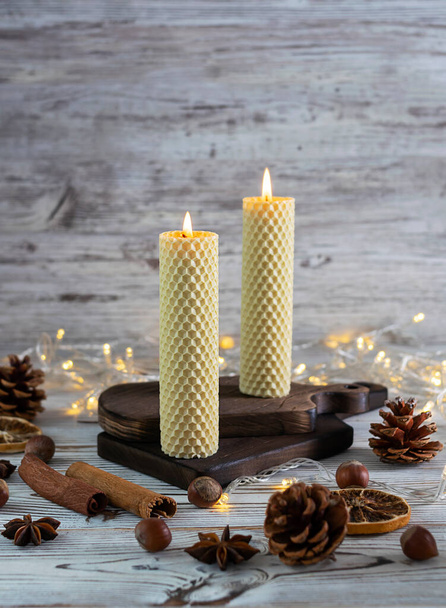 Milk candles made of natural wax stand on a light wooden table. Tubes of cinnamon, stars of star anise and cones in the foreground. Glowing garlands in the background. Light wooden background - Photo, Image
