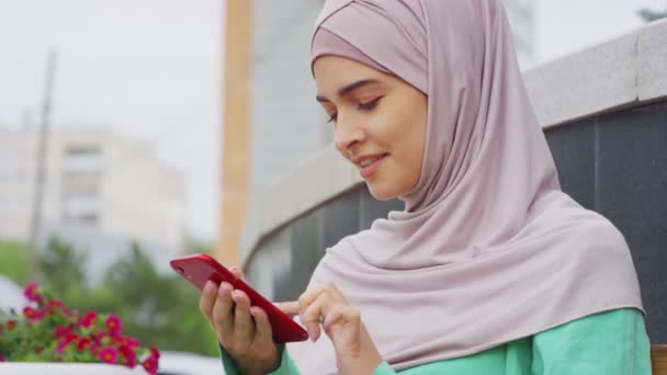 Pecho-up de mujer musulmana joven usando hijab, sentado al aire libre, mensajes de texto en el teléfono inteligente durante el día, sonriendo - Imágenes, Vídeo