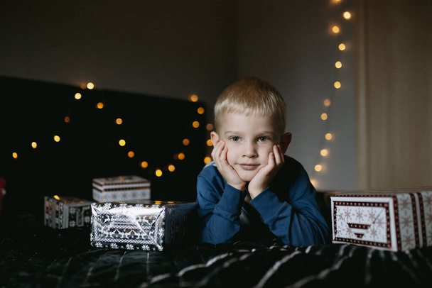 cute little caucasian boy wearing pajamas lying on bed surrounded with christmas presents waiting for holiday. High quality photo - Φωτογραφία, εικόνα