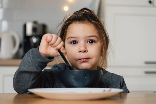 Beautiful child girl eats soup from black bowl with bread and onion. Lifestyle photo of kid in kitchen having a meal. - Foto, Bild