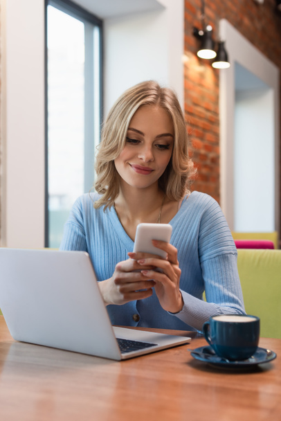 cheerful woman chatting on smartphone near laptop and cup of coffee - Фото, зображення