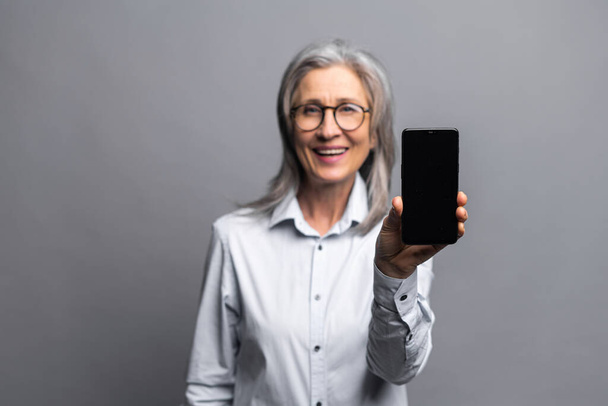 Promotion concept. Pleasant senior gray-haired business woman in glasses and formal wear holding and showing smartphone with empty screen standing on gray, mature lady advertising new app - Photo, image