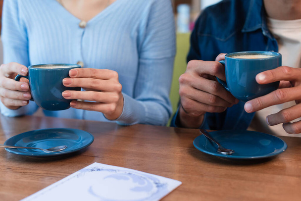 przycięty widok rozmytej pary trzymającej filiżanki kawy w pobliżu menu na stole restauracyjnym - Zdjęcie, obraz