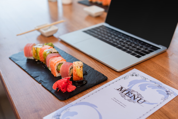 deliciosos rollos de sushi, menú y computadora portátil borrosa con pantalla en blanco en la mesa en el bar de sushi - Foto, imagen
