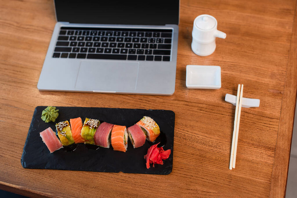 high angle view of plate with sushi rolls, chopsticks, soy sauce pot and laptop with blank screen on wooden table - Photo, Image