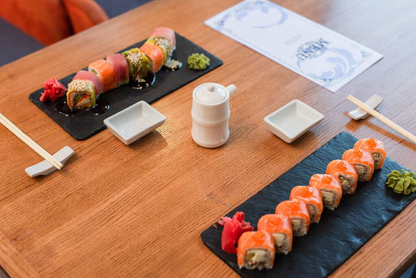 platos negros con deliciosos rollos de sushi con wasabi y jengibre cerca de la olla de salsa de soja y el folleto del menú en la mesa de madera - Foto, imagen