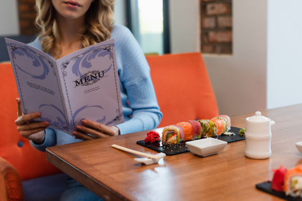 cropped view of blurred woman reading menu near tasty sushi rolls on table - Photo, Image