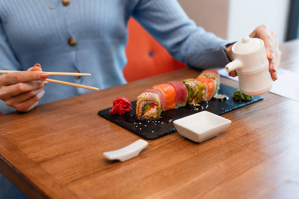 vista parcial de mujer borrosa sosteniendo palillos y olla de salsa de soja cerca de deliciosos rollos de sushi - Foto, Imagen