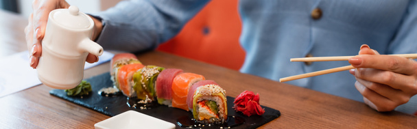cropped view of blurred woman holding chopsticks and soy sauce pot near sushi rolls, banner - Foto, imagen