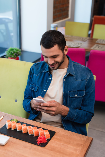 smiling man messaging on mobile phone near delicious sushi in restaurant - Photo, image
