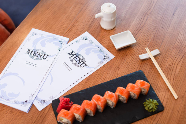 menu brožury v blízkosti lahodné sushi závitky, hůlky, sójová omáčka hrnce a mísa na dřevěném stole - Fotografie, Obrázek