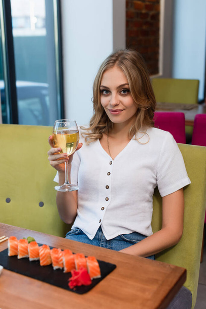 mujer feliz con copa de vino blanco mirando a la cámara cerca de rollos de sushi borrosa - Foto, Imagen