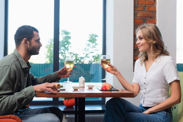 vista lateral de feliz pareja joven sosteniendo vasos de vino blanco durante la cena en sushi bar - Foto, Imagen