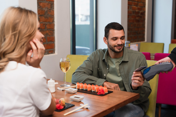 blurred woman near boyfriend paying for dinner in sushi bar through credit card reader - Foto, immagini