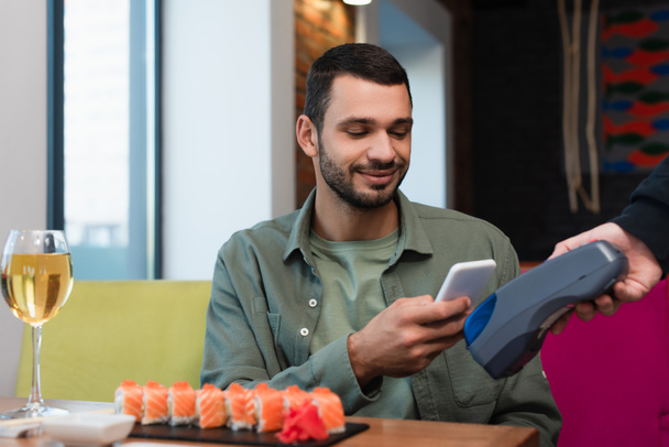 joven pagando con smartphone a través de terminal de pago para sabroso sushi y copa de vino blanco - Foto, Imagen
