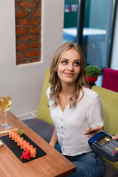 smiling woman with credit card paying through terminal for sushi rolls and glass of wine - Photo, Image