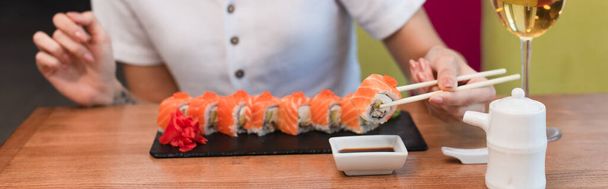partial view of woman with chopsticks near sushi rolls, soy sauce and glass of white wine, banner - Photo, Image