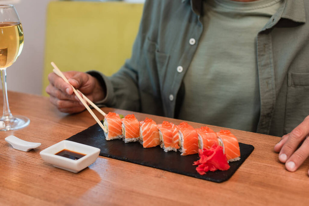 vista recortada del hombre con palillos cerca de sushi delicioso, tazón con salsa de soja y vaso de vino tinto - Foto, Imagen