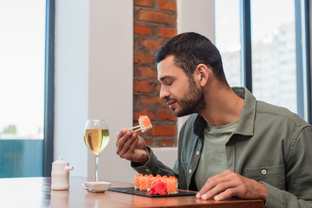 młody człowiek z zamkniętymi oczami trzyma pyszne sushi z pałeczkami w pobliżu szklanki lub białego wina - Zdjęcie, obraz