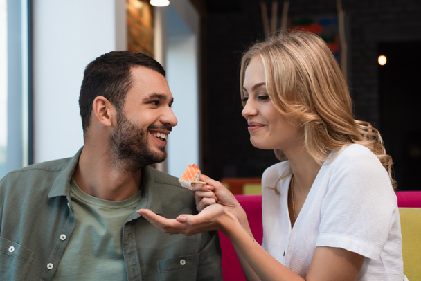 cheerful woman feeding boyfriend with sushi roll during romantic date in restaurant - Foto, Imagem