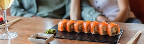 vista recortada de pareja borrosa cerca de conjunto de delicioso sushi, tazón de salsa de soja y vaso de vino blanco, pancarta - Foto, Imagen