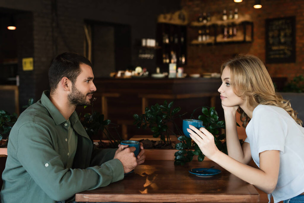 side view of young couple holding coffee cups and looking at each other in cafe - 写真・画像