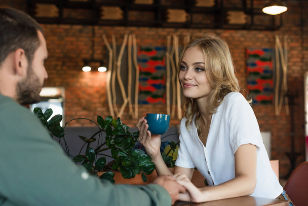 pleased blonde woman with coffee cup holding hands with blurred boyfriend in cafe - Fotó, kép