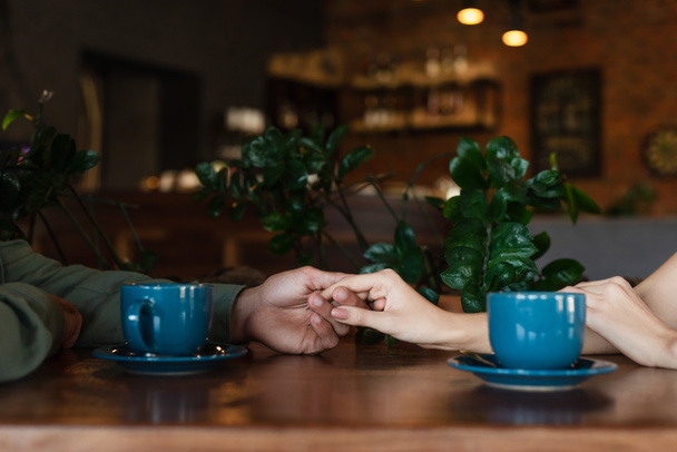 częściowy widok pary trzymającej się za ręce przy filiżance kawy podczas romantycznej randki w kawiarni - Zdjęcie, obraz