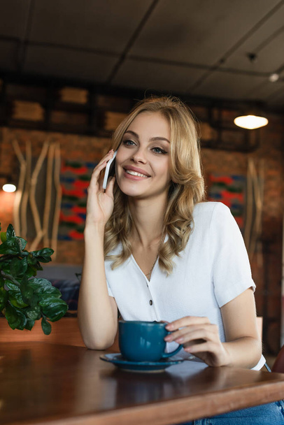 cheerful blonde woman talking on mobile phone near cup of coffee - Fotoğraf, Görsel