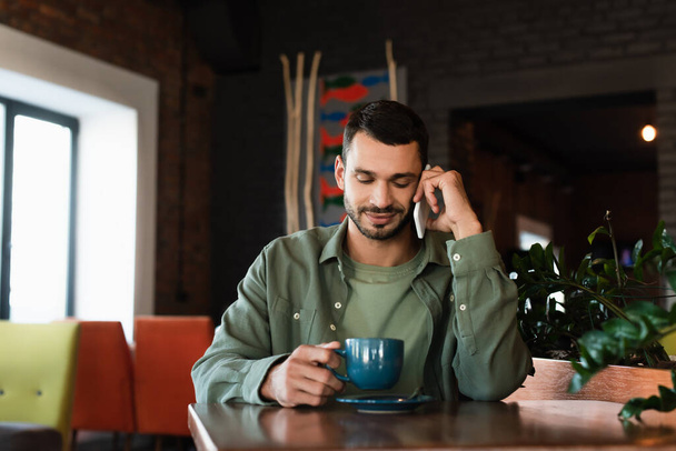 hymyilevä mies pitämällä kuppi kahvia puhuessaan matkapuhelin kahvilassa - Valokuva, kuva