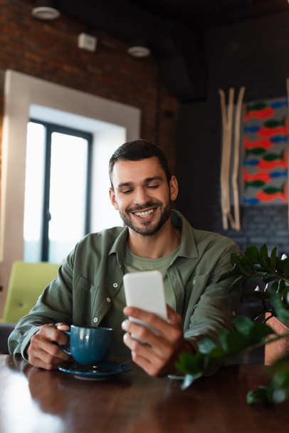 happy man looking at mobile phone while holding coffee cup in restaurant - Фото, изображение