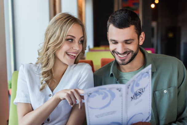 cheerful woman pointing with finger near smiling boyfriend choosing meal from menu - Фото, изображение