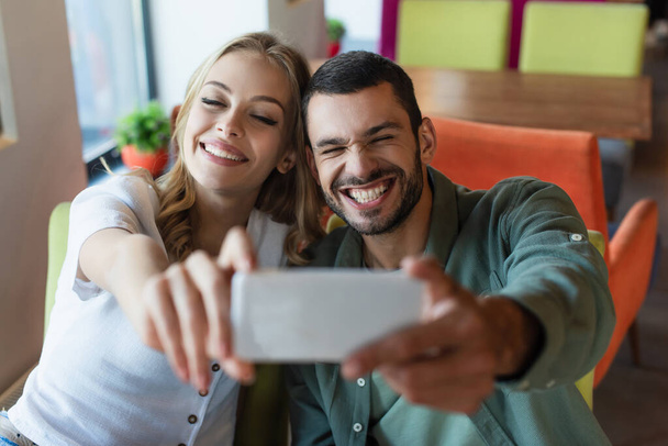 vzrušený muž šklebí poblíž veselá žena, zatímco se selfie na rozmazaný smartphone - Fotografie, Obrázek