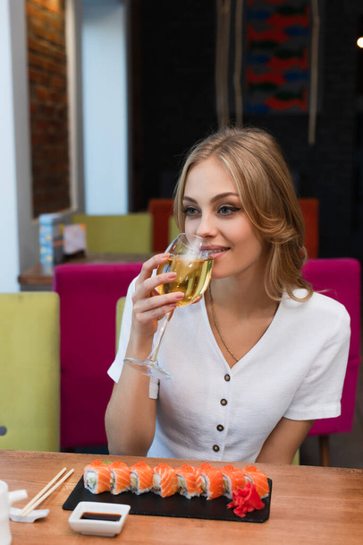 mujer joven y feliz bebiendo vino blanco cerca de sabrosos rollos de sushi - Foto, Imagen