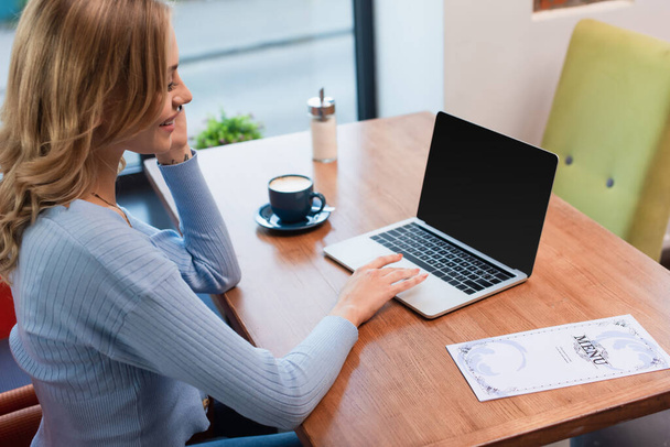 счастливая женщина сидит возле ноутбука с чистым экраном, чашки кофе и меню на столе - Фото, изображение