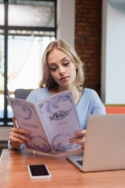 jeune femme blonde lecture menu près d'un ordinateur portable et smartphone avec écran vierge - Photo, image