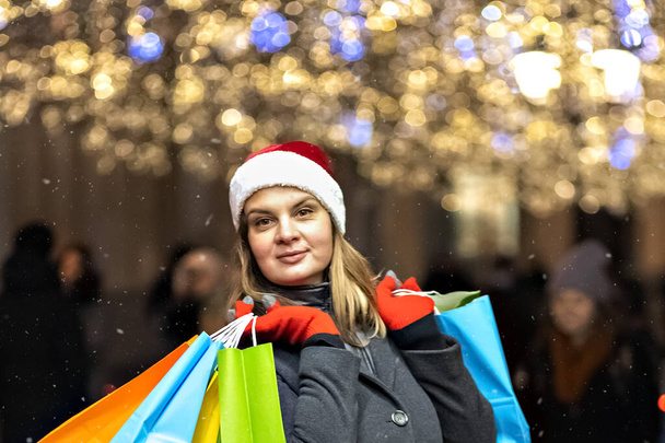 Τα Χριστουγεννιάτικα ψώνια μιας νεαρής γυναίκας. Πολύχρωμο φωτεινό χάρτινες σακούλες με δώρα. Μια βόλτα στα χριστουγεννιάτικα μαγαζιά μιας νεαρής όμορφης γυναίκας. Γιορτάζουμε την Πρωτοχρονιά.. - Φωτογραφία, εικόνα