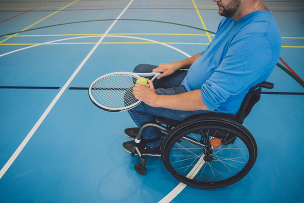 Взрослый человек с ограниченными физическими возможностями, который использует инвалидное кресло, играя в теннис на крытом теннисном корте - Фото, изображение
