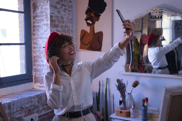 Genç bir kadın, ressam, tasarımcı ve kırmızı bereli bir sanatçı, cep telefonuyla stüdyosunun içinde selfie çekiyor. Konsept fotoğraf, fotoğraf, selfie, cep telefonu. - Fotoğraf, Görsel