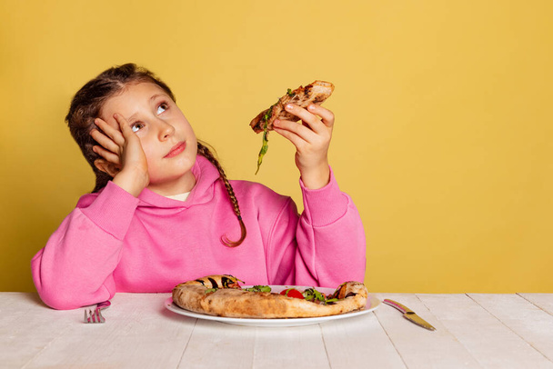 Studio shot of little cute girl sitting and tasting delicious Italian pizza isolated on yellow studio background. - Фото, изображение