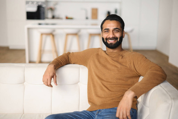 Satisfied young Indian man resting on comfortable couch in white cozy modern living room, looking at the camera with toothy smile. Millennial hipster guy enjoy no stress life - Foto, Bild