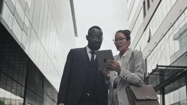 Niski kąt śledzenia zdjęć Afroamerykańskiego biznesmena i azjatyckiej biznesmenki w formalnym ubraniu stojącej na ulicy w mieście i dyskutującej o pracy patrząc na tablet - Materiał filmowy, wideo